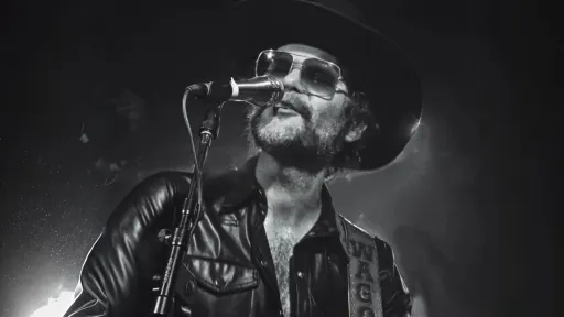 Black and white photo of man wearing a leather jacket, sunglasses and a cowboy hat standing at a microphone playing the guitar.