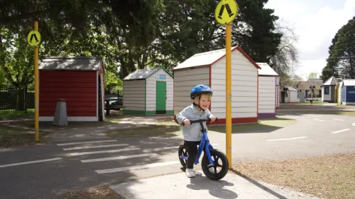 Child wearing a denim jacket and blue helmet rides a balance bike across a zebra crossing. In the background are small buildings.