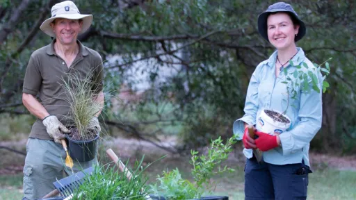Two people in a park holding potted plants with a wheelbarrow full of plants