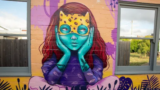 A mural of a child sitting cross legged with their chin in their hands. They have red flowing hair and are wearing a cat mask over their eyes.