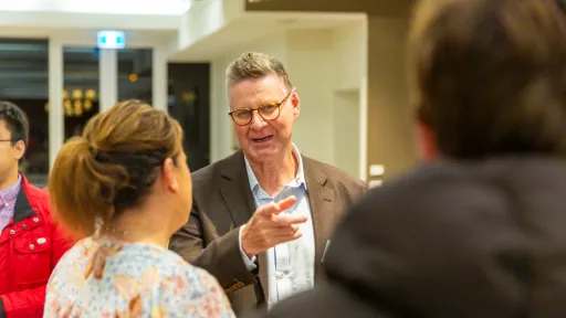 Man with glasses wearing a brown jacket and blue shirt talking to a lady with brown hair in a pony tail wearing a floral blouse.