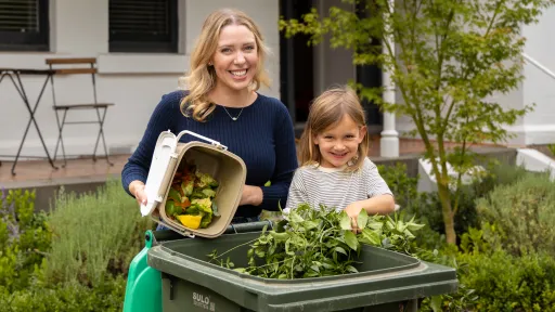 Woman and child tip food scraps and branches into bin with green lid