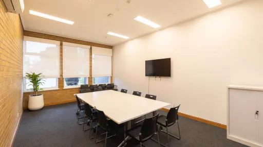A meeting room with a table surrounded by black chairs. The room has a TV. 
