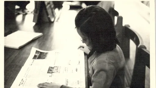 A black and white image shows a young child sitting at a wooden desk reading a newspaper