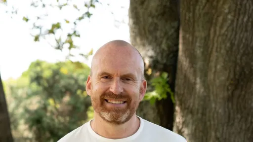 A person with a bald head and a beard is smiling while standing in front of a tree