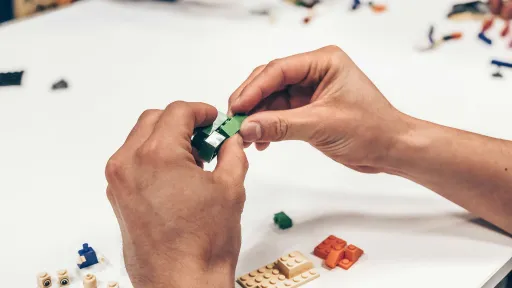 Two hands are joining together pieces of lego. There is also lego pieces sitting on a table nearby.