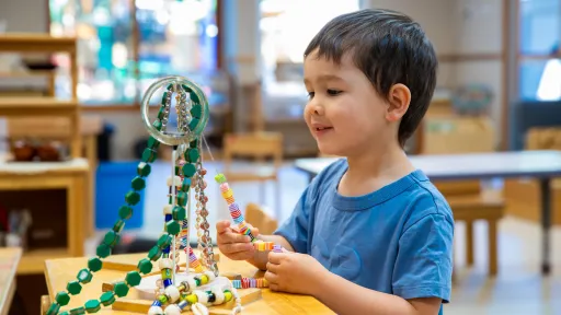 A jewellery stand is on a table in front of a child. The child is practicing threading beads onto string.