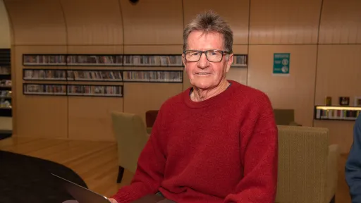 Man in red jumper sits with laptop and library shelves in background