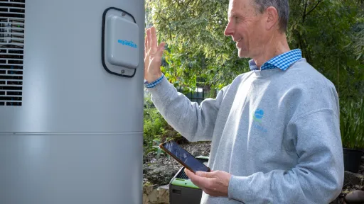 Man in grey jumper stands next to tall hot water cylinder in yard