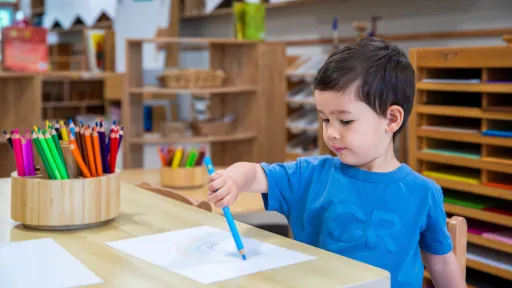 Child sitting at a table drawing a rainbow. They're holding a blue pencil.