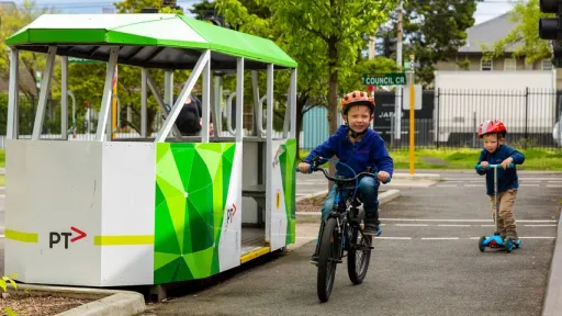 Two children, one on a bike, the other on a scooter riding along a path that bypasses a model tram at Kew Traffic School.
