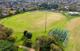 Aerial view of oval sportsground with various square line markings on its surface and four tall light posts around its edge. There is a pavilion to the top right and cricket nets to the bottom right.