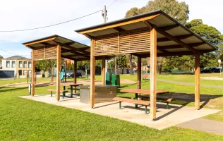 Two under cover picnic tables with a bbq between the two. There is a concrete path leading to the undercover area. A playground and residential houses can be seen in the background. 
