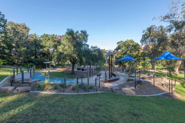 Man-made garden with a tall brown sculpture, rock feature and two blue umbrellas to the right. It includes concrete and tan bark sections and is surrounced by grass. There are trees in the background.