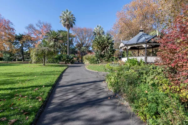 Concrete path with grass areas on either side. There is a covered rotunda with lattice patterns to the right, and tall trees in the distance.