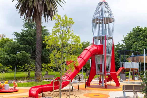 Playground with rocket-shaped climbing feature and slide, and small carousel and park bench. There are several trees in the playing area and the background.