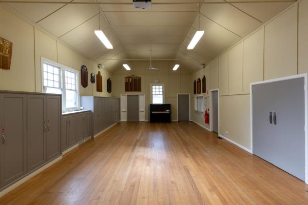 Inside of a hall with polished floorboards and a piano at the far end. There are two grey doors on the right and grey cupboards and a window along the left.