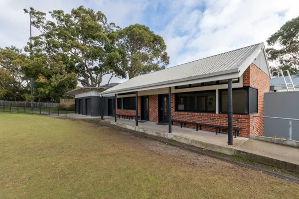Single-storey brown brick building with triangular roof and black window frames. There are tall trees in the background.