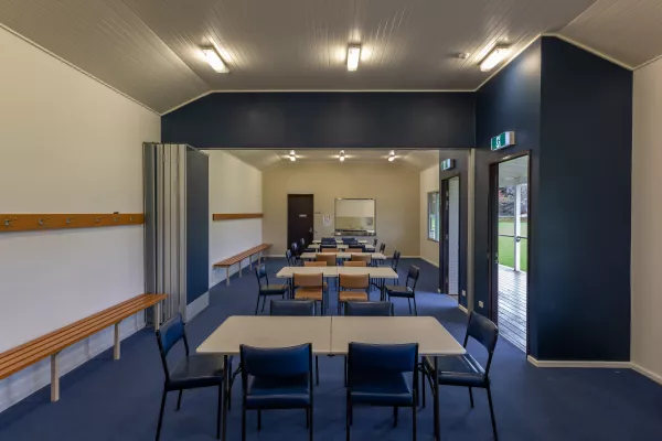 Long thin room with six white tables and 30 blue and brown chairs. There are coat racks and bench seats along the walls, two doors to right and a kitchen at the far end. The lights are switched on.