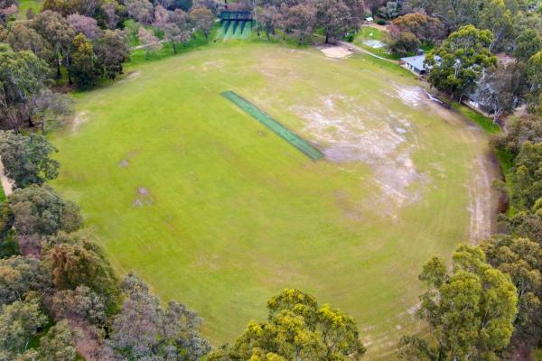 Aerial view of oval field with cricket pitch and some bare patches, surrounded by a thick layer of trees. There are cricket nets at the top and a small building to the top right.
