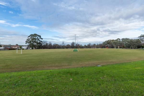 Oval sportsground with soccer goals at each end and a dark green cricket pitch in the middle. There are trees and three light posts on the horizon.