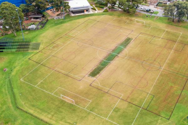 Aerial view of sports field with white markings for soccer and black markings for another sport. There are cricket practice nets to the left, a pavilion at the top and a road to the top right.