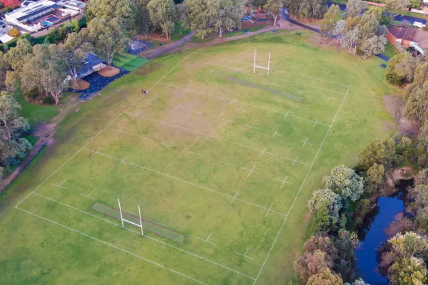 Aerial view of playground to the south and sportsground with rugby goal posts to the north. There are walking paths on each side of the playground and thick mature trees surround the area.