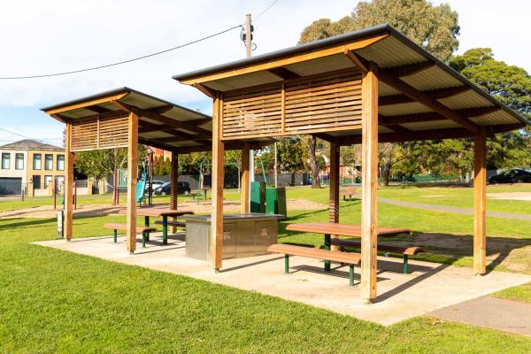 Barbecue area in a park, with 2 sets of tables and chairs and 2 wooden shelter structures with diagonal roofing.