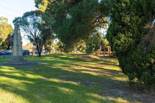 Small grass area with tall concrete memorial structure to the left and tree shadows throughout. There is a thick tree and bare dirt patches to the near right. A road and cars are visible to the left.