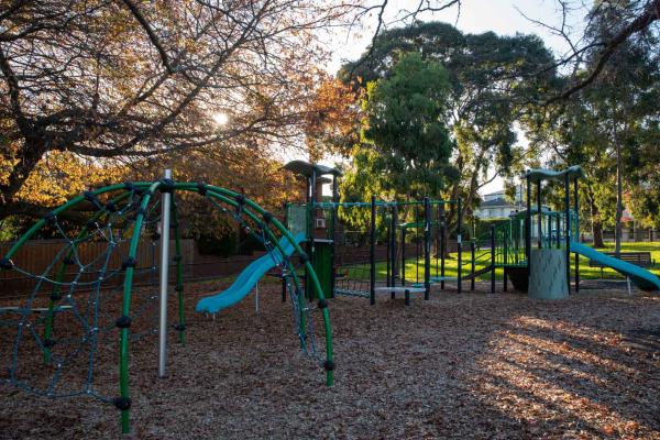 Playground in shadows with large climbing feature, two slides and a dome-shaped feature. There are tall trees and large houses in the background, with sunlight coming through.
