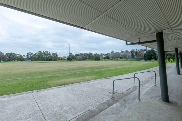 Looking out towards a green sports field from a sports grandstand . Trees and houses are visible in the background. 