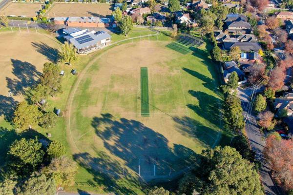 Aerial view of an oval sportsgrouind. A green cricket pitch is in the centre. The oval is surrounded by houses and trees. 