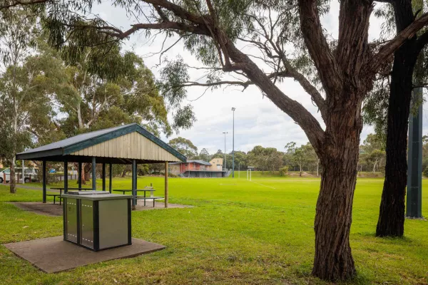 A park area with a barbecue and an undercover seating area. A sportsfield is in the background.