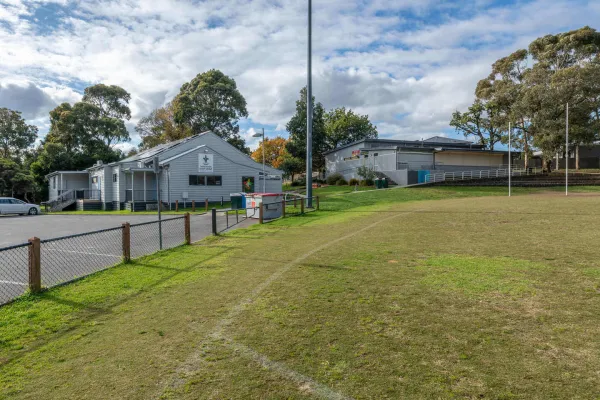 The edge of a sportsground oval with line markings and a short wire fence. 2 buildings are next to the oval. 
