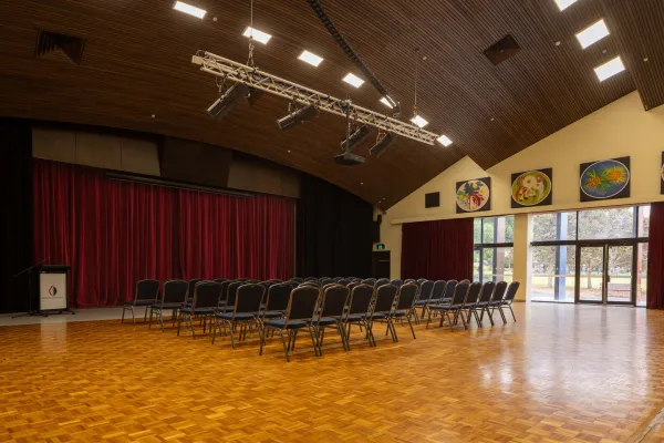 A room with rows of chairs facing a stage area with red curtains. Glass doors look out into a green area. The room has a wooden floor