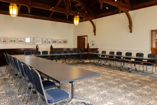 A large room with with long tables arranged in a U-shape. The room features dark wood ceiling, hanging lantern-style lights, and patterned carpet. 