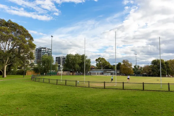 AFL oval with white goalposts, surrounded by a chain-link fence. Two people are running on the field. Trees line the edge, and apartment buildings are visible. 