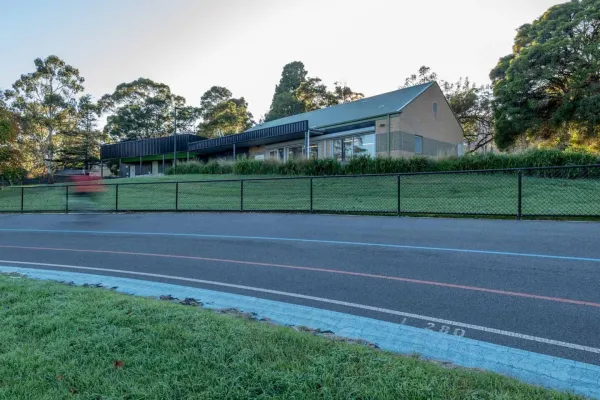 A velodrome track with blue and red line markets. In the background is short wire fence, and a building surrounded by trees.