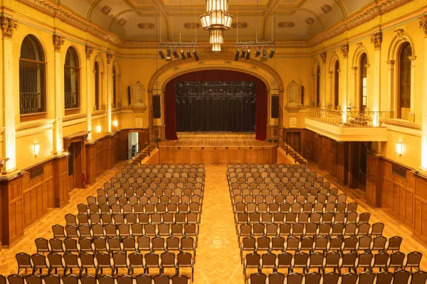 The spacious auditorium's interior includes a stage. Rows of chairs are set up in the main floor. There is a balcony on the right side and a large stage with red curtains at the far end. The room has ornate architectural details and is lit with chandeliers.