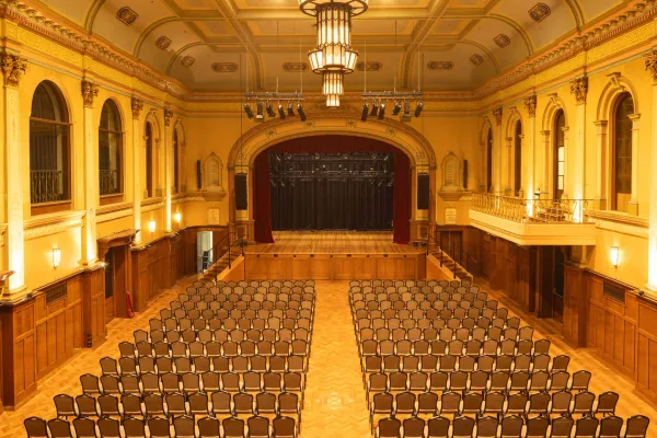 The spacious auditorium's interior includes a stage. Rows of chairs are set up in the main floor. There is a balcony on the right side and a large stage with red curtains at the far end. The room has ornate architectural details and is lit with chandeliers.