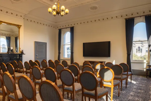 A meeting room featuring rows of wooden chairs with gray fabric seats, arranged in front of a large, wall-mounted television. Two windows flank the screen, draped with dark curtains. An ornate fireplace and mirror are visible to the left. The room has detailed ceiling molding and a chandelier.