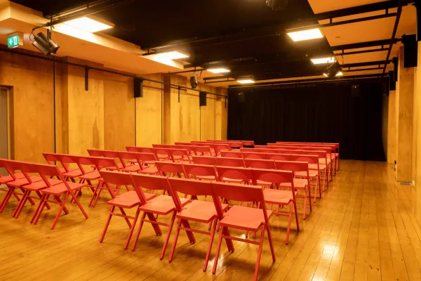 A small event space with rows of red folding chairs facing a black curtained stage. The walls are wood paneled and there are lighting fixtures and speakers mounted on black tracks on the ceiling. 