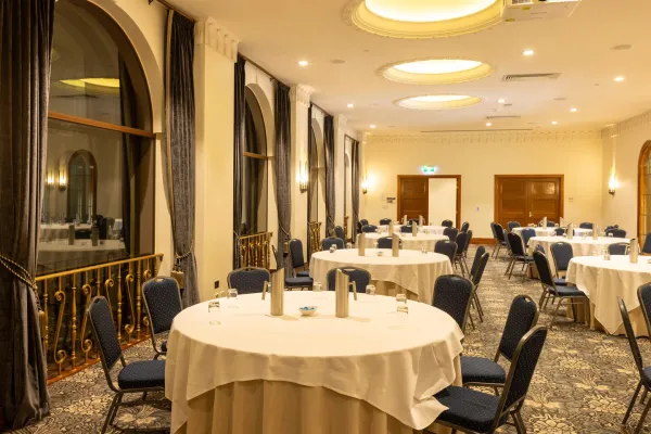 A conference room is set up with round tables covered in white cloths, each surrounded by chairs with blue patterned upholstery. There are large, arched windows with dark curtains along one side of the room. The ceiling features recessed lighting and a projector. The floor is covered with a patterned carpet.