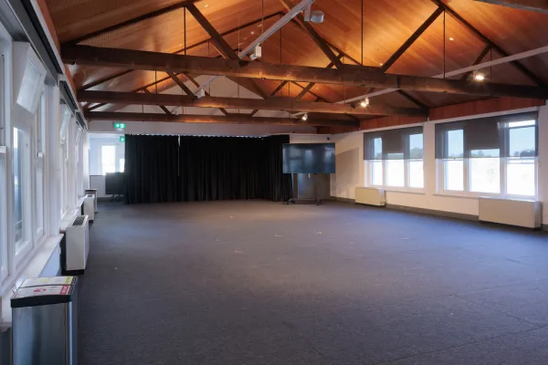 Wide shot of an empty event space with a high, angled wooden ceiling supported by exposed beams. Dark gray carpet covers the floor. A wall of windows is on the left, and another wall of windows with dark blinds is on the right. Black curtains are draped on the far wall, with a large flat-screen TV in front.