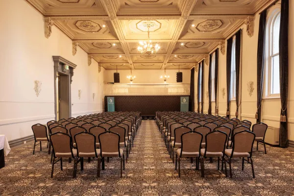 An interior view of a large room set up for an event. Rows of chairs face the back of the room, where a raised platform is centered.  The ceiling is ornately decorated with square recessed panels and gold trim and chandeliers  Tall windows line one side of the room, draped with dark curtains. The floor is covered with a patterned carpet.