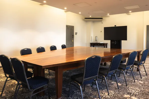 A long rectangular wooden table is surrounded by blue patterned chairs in the meeting room. A large screen is mounted on the wall at one end of the table.