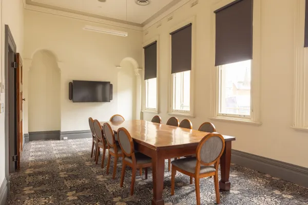 A meeting room featuring a long rectangular wooden table with 10 chairs. A TV sits on the far wall. There are tall windows and patterned carpet. 
