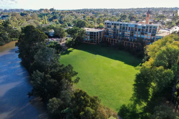 Birds eye view of a large square park space next to a river. The park is surrounded by trees and a apartment building