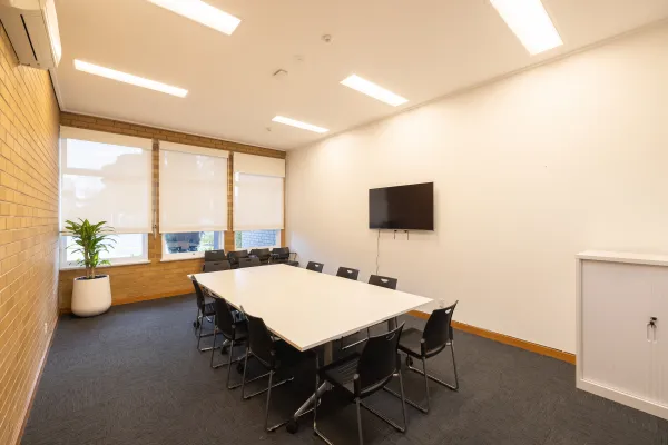 A meeting room with a table surrounded by black chairs. The room has a TV. 