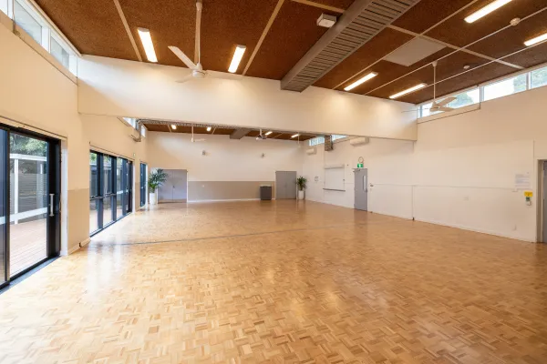 A large hall with wooden floors. There are windows on the left looking out to a decked area. 